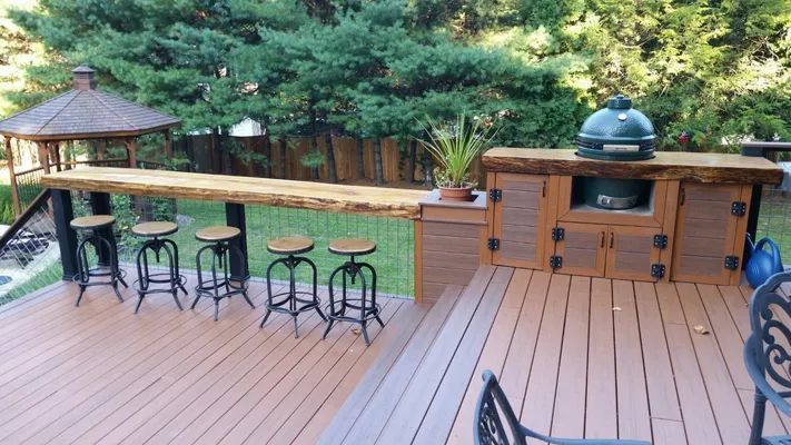 Outdoor deck with built-in grill and bar. Brown wooden deck and furniture, lush greenery in background.