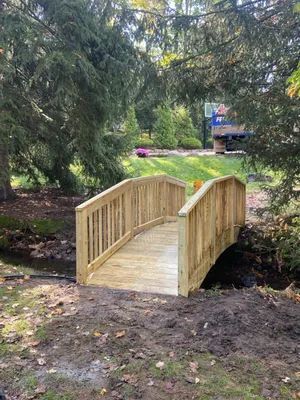 Wooden arched bridge spans a small stream in a wooded area.