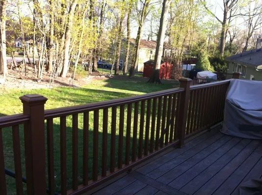 Wooden deck with brown railing overlooking a grassy yard and trees in sunlight.