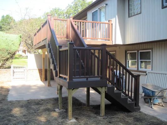 Wooden deck and stairs attached to a two-story house, painted brown. The ground is dirt and concrete.