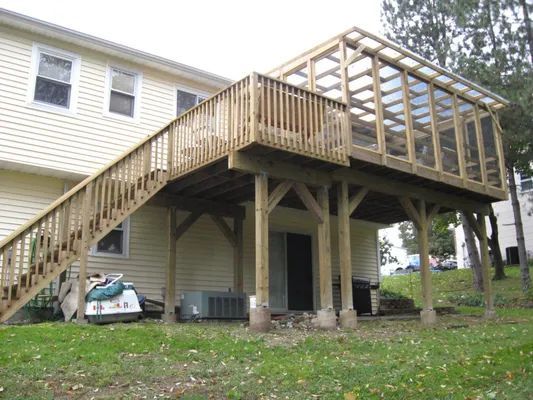 Wooden deck attached to a two-story house with a covered sunroom and staircase, supported by posts.