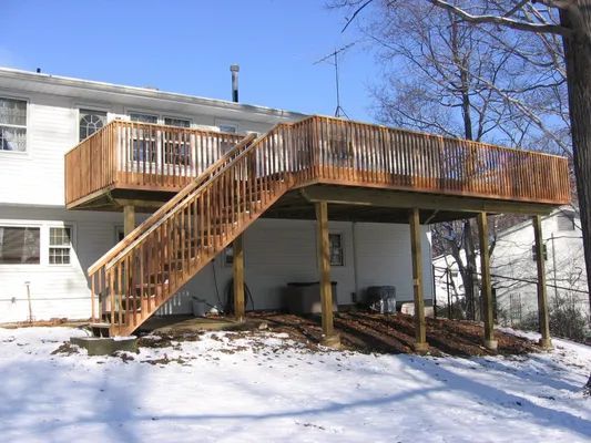 Wooden deck with stairs attached to a two-story house, set in a snowy landscape.