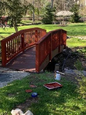 Wooden arched bridge, stained brown, over a dark stream in a grassy yard, paint supplies visible nearby.