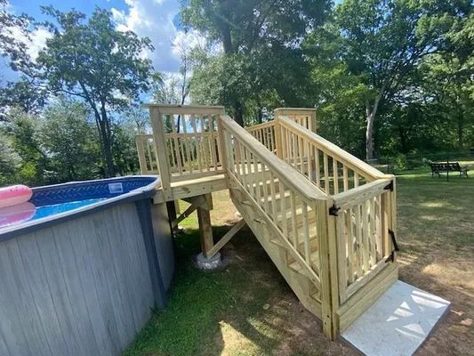 Wooden deck with stairs leading to an above-ground pool. Green trees and grass in the background.