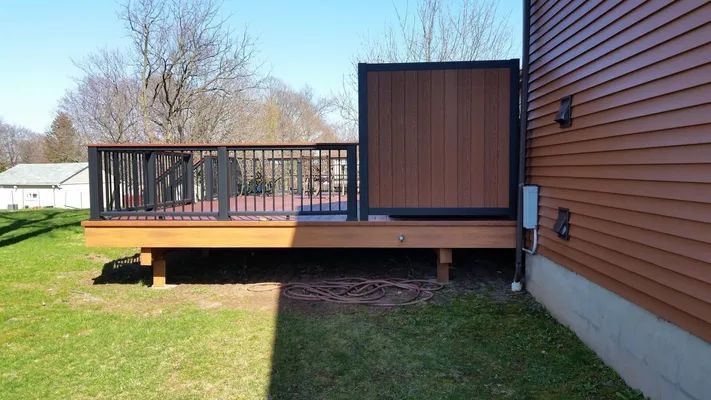 Wooden deck with black railing and brown privacy wall attached to a brown house.