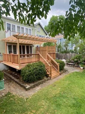 Wooden deck with pergola attached to a two-story green house in a backyard setting.