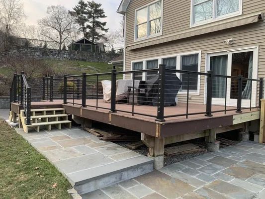 Wooden deck with black railing and steps, leading to a stone patio next to a house.