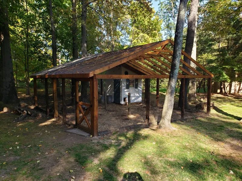Wooden shelter in a forest, brown roof, posts, surrounding trees, and a small white structure inside.