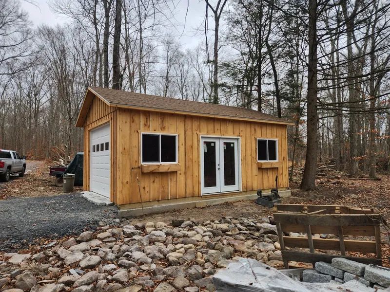 Wooden building with garage door, windows, and double doors; set in a wooded area with gravel and stone.