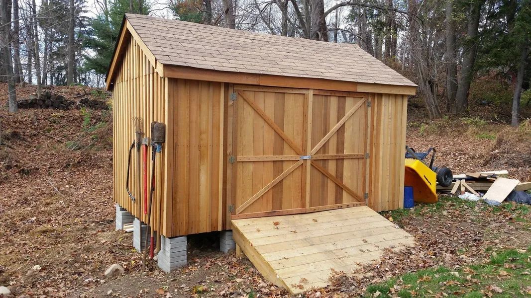 Wooden shed with a ramp and tools leaning on the side, surrounded by foliage.