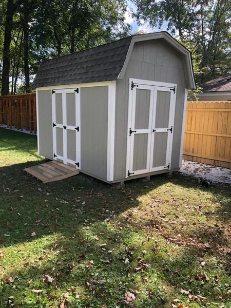 A small gray shed with white trim and a brown roof sits on green grass.