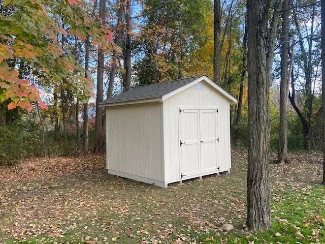 Beige shed with double doors in a wooded area with fall foliage.
