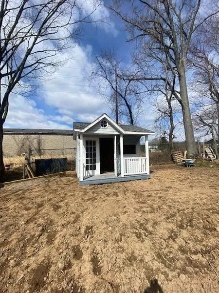 Small white shed with gray trim and porch, set on a dirt patch surrounded by trees under a partly cloudy sky.