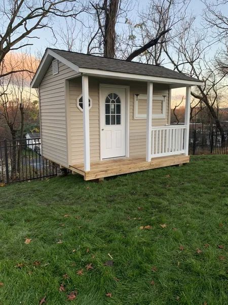Small beige shed with porch and white trim on grassy hill.