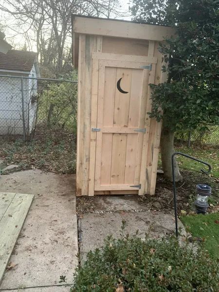Wooden outhouse with a crescent moon cutout on the door, sitting outdoors.