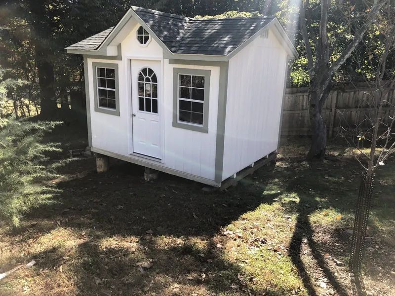 White shed with green trim and gray roof in a grassy yard, surrounded by trees.