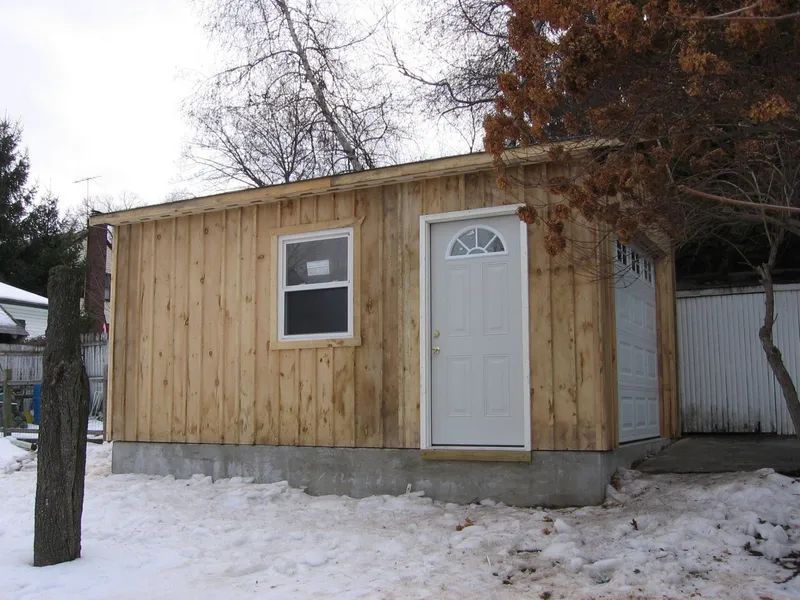 Wooden shed with white door, window, and garage door on a concrete foundation in snow.