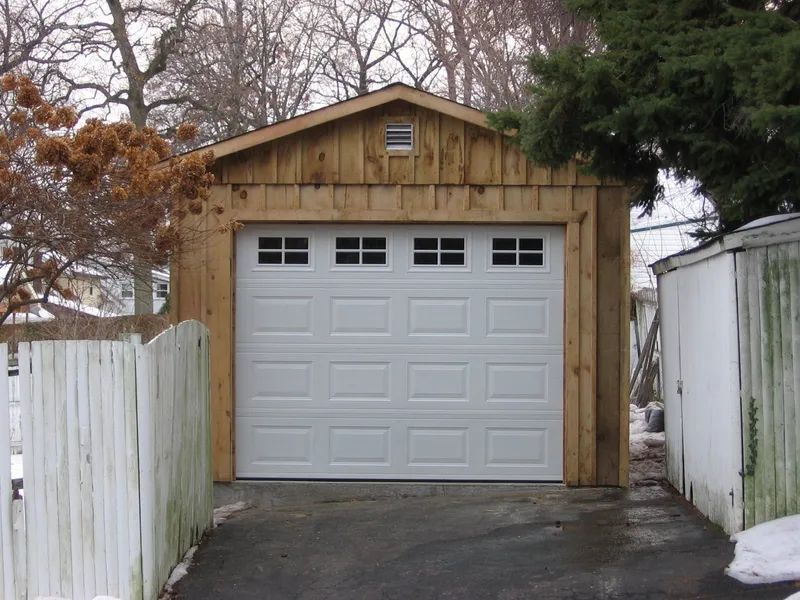White garage door on a wooden garage with a peaked roof; a driveway and fence are in front.
