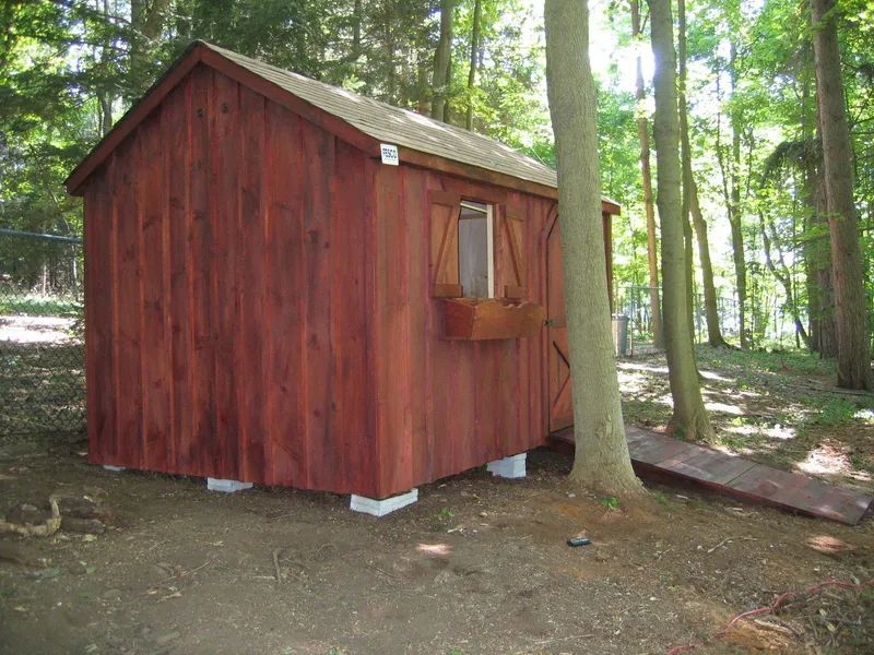 Red wooden shed on concrete blocks, with a window and ramp in a wooded area.
