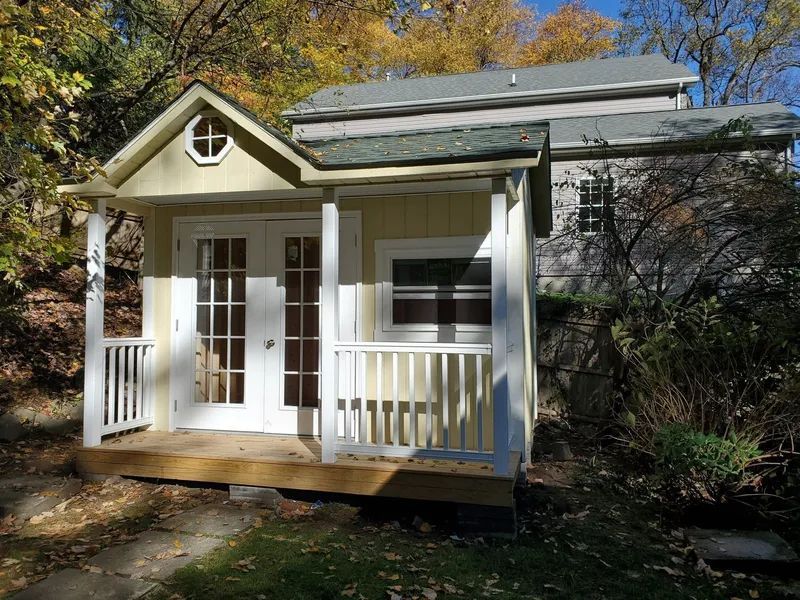 Yellow shed with porch and white doors, set against a two-story building in a wooded area.