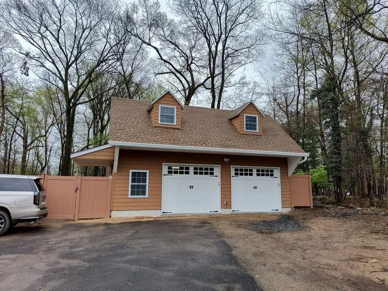 Two-car brown garage with white doors, dormers, and a brown fence. A white SUV is parked to the left.