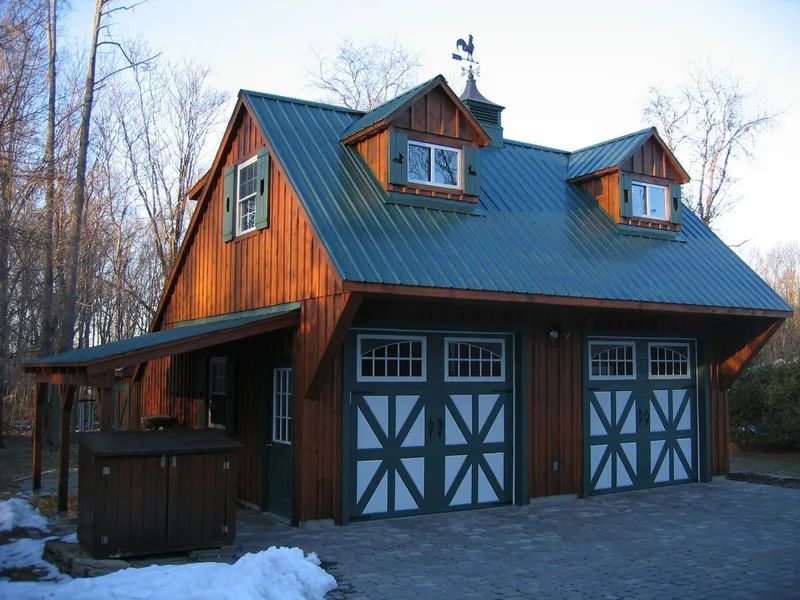Two-car garage with green metal roof, dormers, and brown wooden exterior; a small porch is to the left.