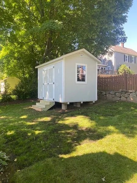 Pale blue shed with white trim and a small window, sits on concrete blocks. Wooden steps lead up to a double door.