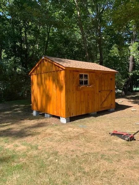 Wooden shed on concrete blocks, stained orange-brown. Small window and door visible, set in a grassy yard.