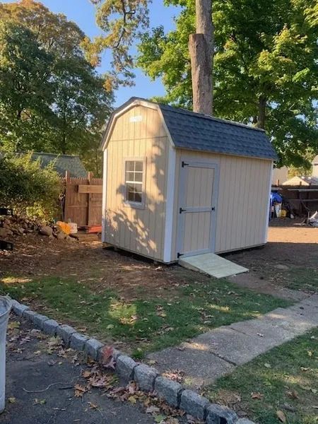 Tan shed with a black roof, a small window, and a wooden ramp, on a grassy patch.