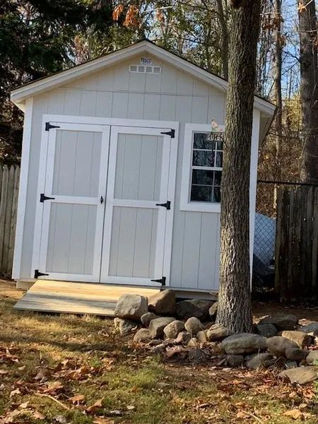 Gray shed with double doors, small window, and a wooden base, next to a tree, in a yard.