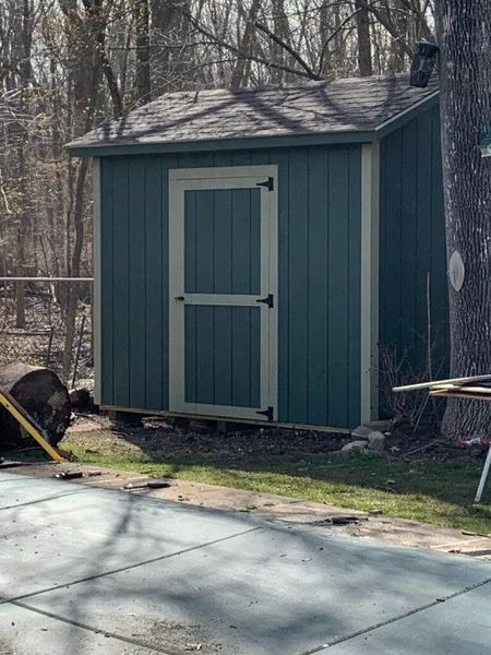Blue shed with off-white trim, gray roof, black door hardware, set in a yard.