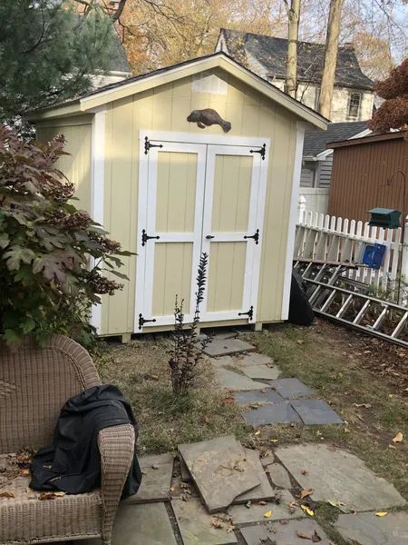Yellow storage shed with white-trimmed double doors and stone path in a backyard setting.