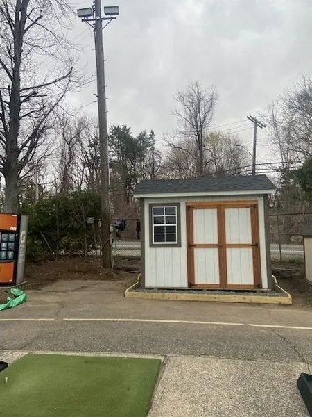 Small white shed with brown doors and trim, next to a concrete area and utility pole, under a cloudy sky.
