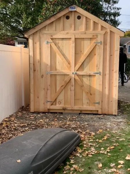 Wooden shed with double doors, standing on gravel. Leaves and a car top carrier are in front.