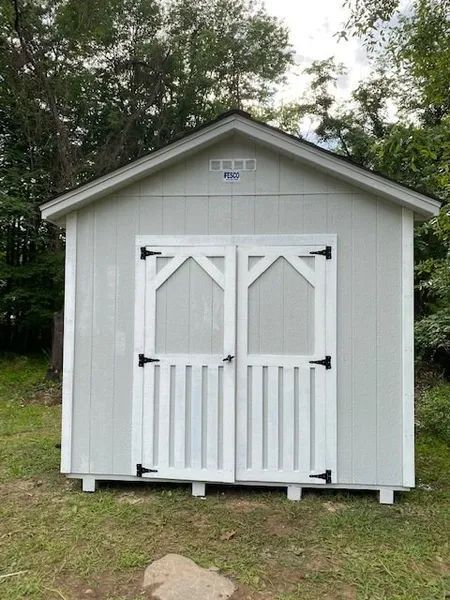 Gray shed with white doors in a grassy yard, surrounded by trees.