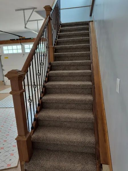 Staircase with brown carpet, wooden railing, and black wrought iron balusters. The stairs lead upwards.