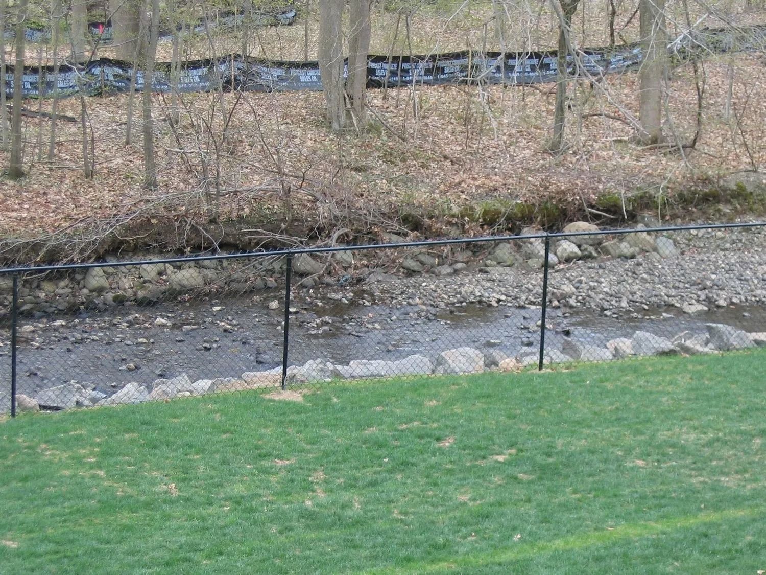 Black chain-link fence bordering a rocky stream, with trees and grass in the background.