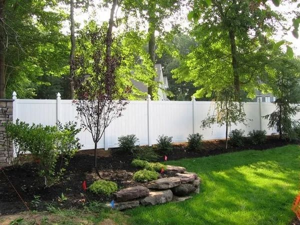 White fence in a sunny backyard with greenery, trees, and landscaping.
