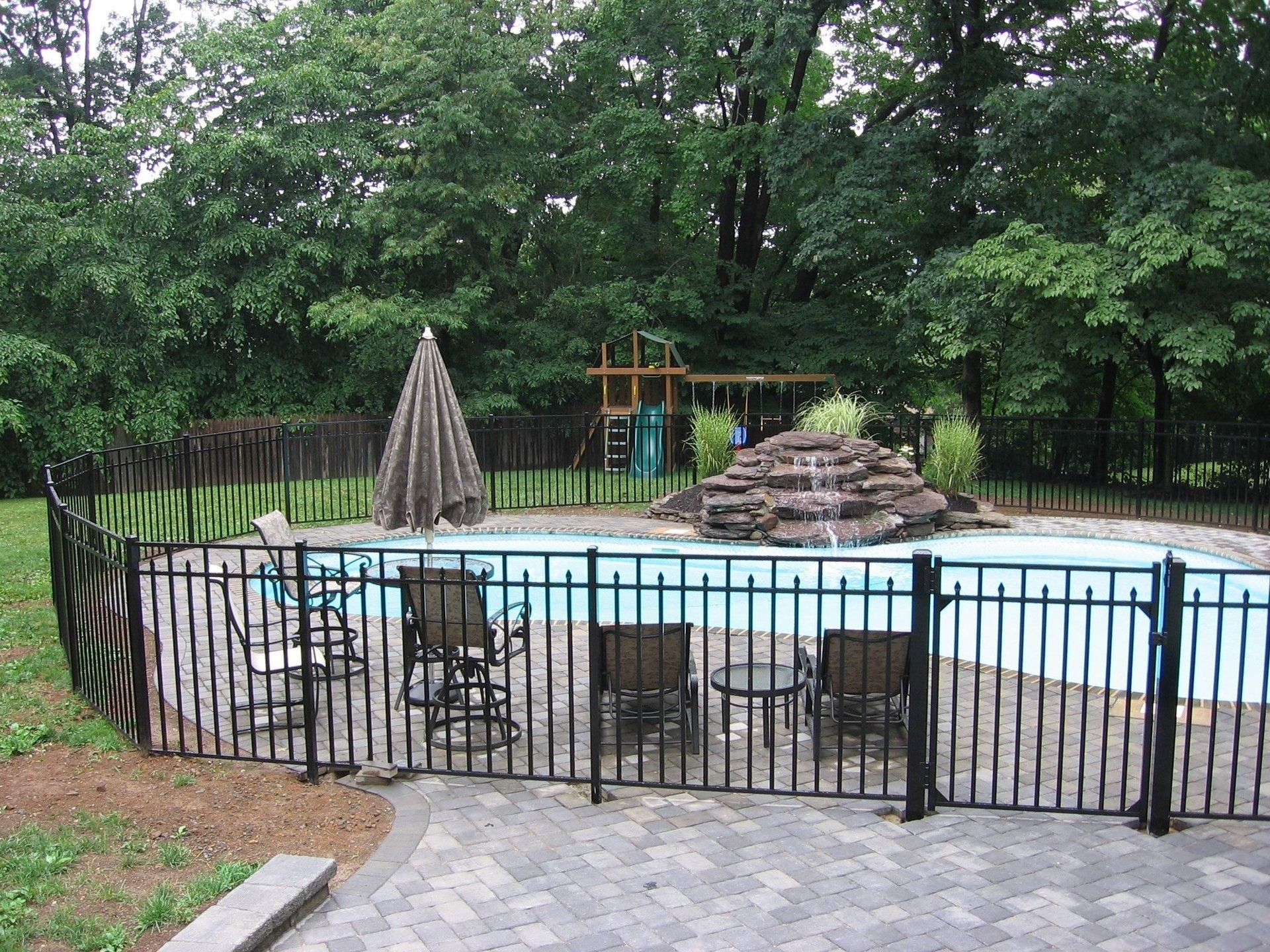 A backyard pool with black fencing, surrounded by trees and a paved patio with chairs and an umbrella.