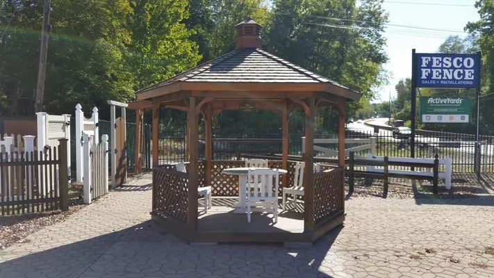 Wooden gazebo with a table and chairs, displayed by a fence company on a sunny day.