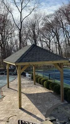 Wooden gazebo with a shingled roof, on a paved patio near a tennis court and trees.