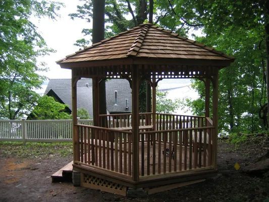Wooden gazebo with shingled roof in a wooded area. The gazebo has railings and sits on a small deck.