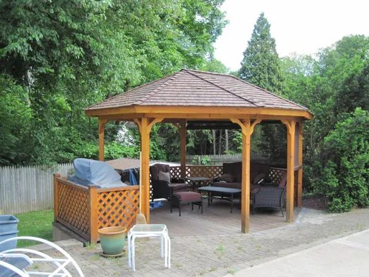 Wooden gazebo with seating on a patio, surrounded by trees and a fence.