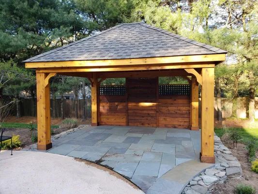 Wooden gazebo with a brown roof and a stone patio, set in a backyard.