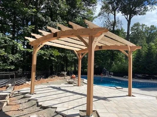 Wooden pergola over a pool, on a stone patio, with trees in the background.