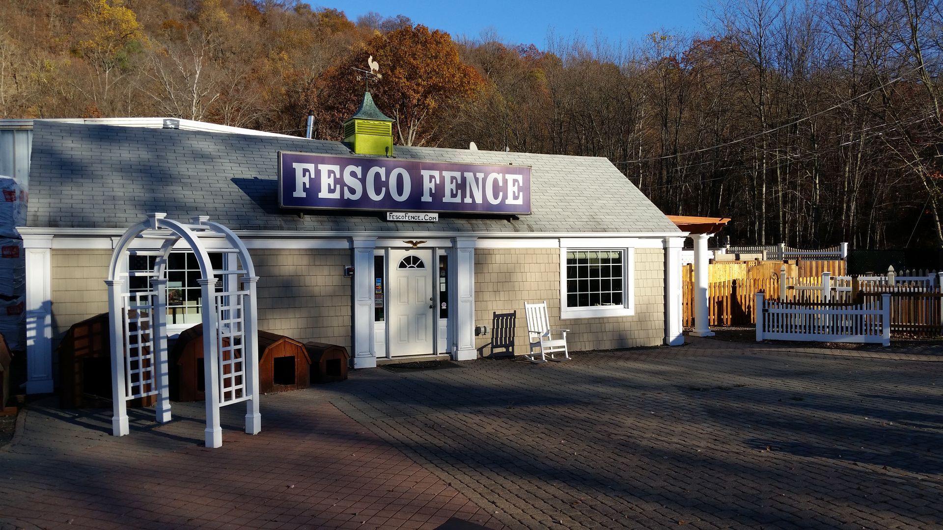 FESCO Fence building with sign, blue door, and small display of outdoor furniture. Background of trees and blue sky.