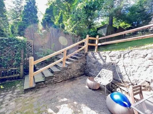 Stone steps with wooden railing lead up to a yard, patio furniture sits below, trees and a fence are in the background.