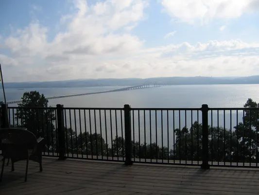View of a bridge spanning a wide lake from a wooden deck with a black railing, cloudy sky.