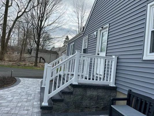 Gray house with white railing and steps. A paved walkway leads to the entrance.