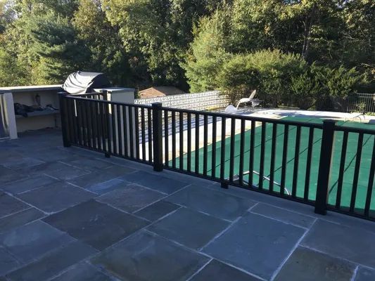 Black fence surrounding a pool on a stone patio. Trees in the background.